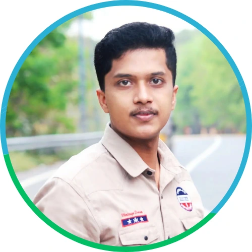 A young man with short black hair and a slight smile, wearing a beige shirt, standing outdoors on a road with greenery in the background.